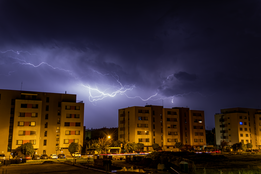 Striking lightning illuminates the night sky above urban apartments, showcasing a dramatic evening scene.